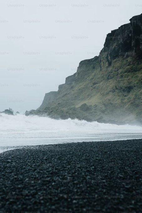 A misty coastal scene shows powerful waves crashing against a dark, moss-covered cliff face, with a black pebble beach in the foreground bearing the "Unsplash+" watermark. The image visually parallels the post's message about the harsh, overwhelming conditions traders face—symbolizing how even when the "chart" (the visible landscape) is clear, the turbulent emotional waves can still pull one under without proper psychological preparation to act on the plan.