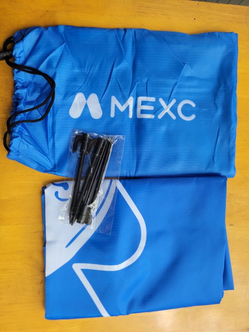 A blue MEXC branded drawstring bag with a small plastic bag containing black plastic items placed on top of it, sitting on a wooden surface.