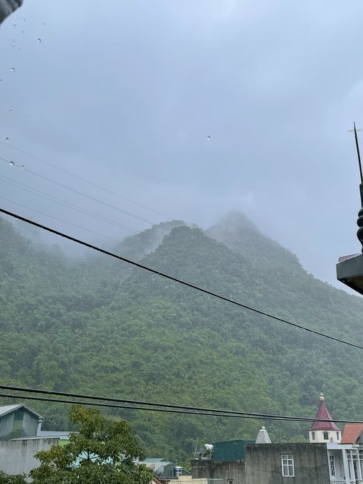 A misty, overcast landscape view of a lush green mountain seen from a town, with power lines crossing the foreground and raindrops visible on the lens.