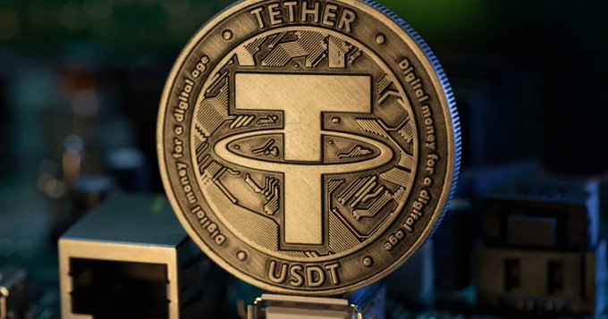 A close-up of a gold-colored Tether (USDT) cryptocurrency coin with the 'T' logo, displayed against a blurred dark background with the text 'Digital money for a digital age' engraved around its edge.