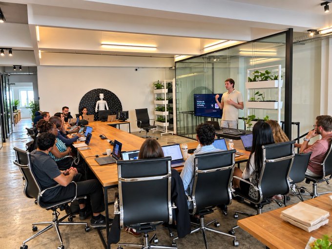 A modern conference room where a man stands at the front presenting to a group of attendees seated around a long wooden table with laptops, with a TV screen displaying a presentation slide behind him.