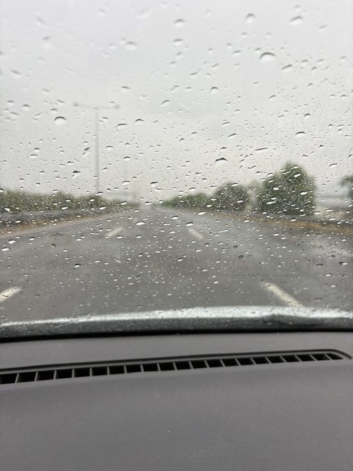 A view from inside a car looking through a rain-covered windshield at an empty parking lot on an overcast day.