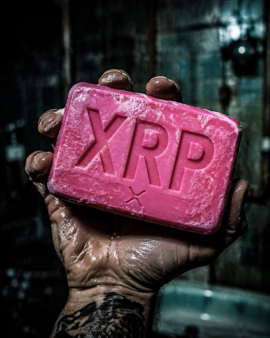 A close-up shows a wet, dirty hand with tattoos holding a pink bar of soap embossed with 'XRP' (referencing the Ripple cryptocurrency) against a dark, gritty background.
