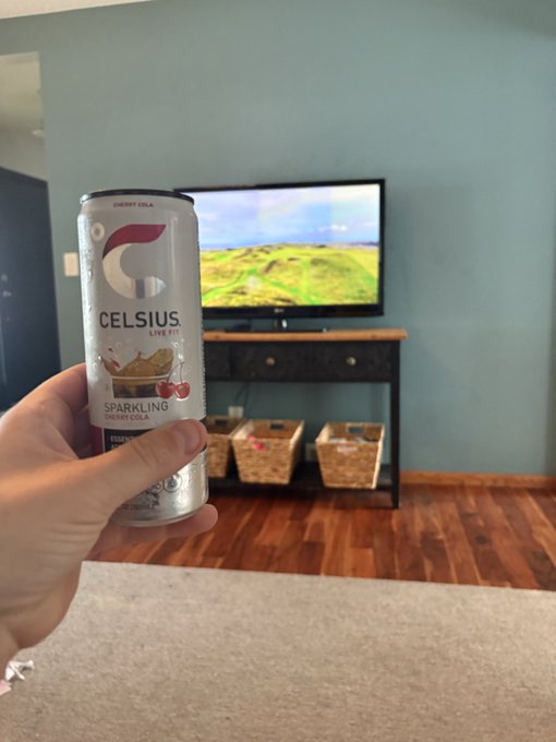 A hand holds a can of CELSIUS Sparkling Cherry Cola in a living room with a TV displaying a golf video game and storage baskets on a console table in the background.