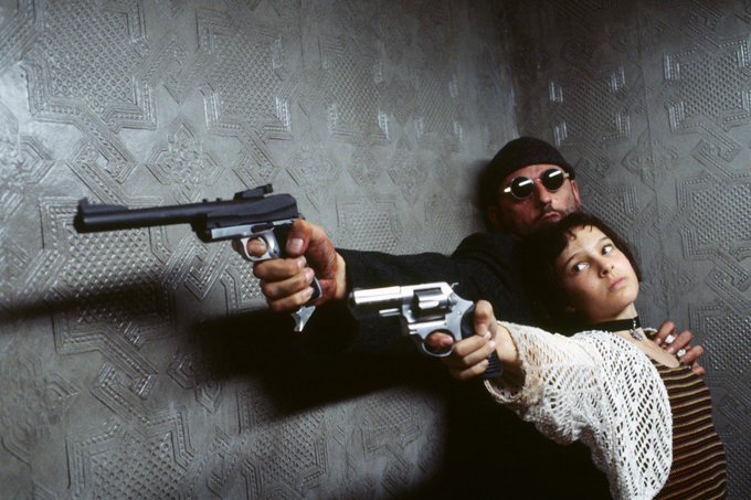 The image shows a man holding a gun, while a woman stands behind him with her arms around his neck. The scene appears to be set against a gray wall or background. There are also two other people present in the image, one of them wearing sunglasses. The main focus is on the interaction between the man and the woman, possibly indicating an intense situation or a dramatic moment captured in time.