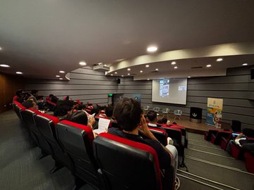 A photograph taken from the back of a lecture hall showing an audience seated in red and black chairs watching a presentation on a projector screen at the front of the room.