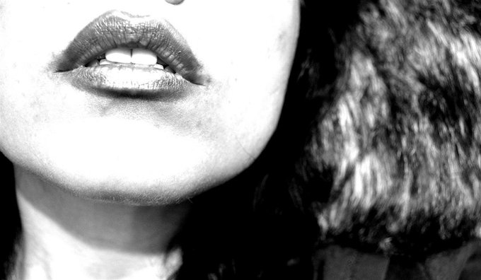 A black and white close-up photograph focusing on a woman's lips, with dark curly hair visible in the background.