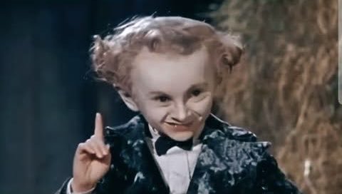 A still image of a character with styled hair buns, dressed in a patterned suit jacket and bow tie, pointing a finger upward with a sly expression.