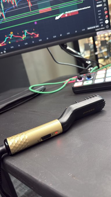 A gold and black hair styling tool branded "champagne" rests on a desk in front of a trading monitor displaying candlestick charts, hinting at a playful misdirection in the post about "shooting the daily entry"—the word "shoot" and the chart context suggest a trading setup, not a camera, despite the presence of what might be mistaken for filming equipment.