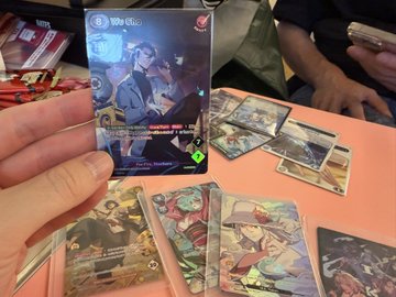 A person holding up a trading card showing an anime-style character with a '3' in the top left corner, with other cards scattered on a table below, including characters with text 'Duelist' and other Japanese text.