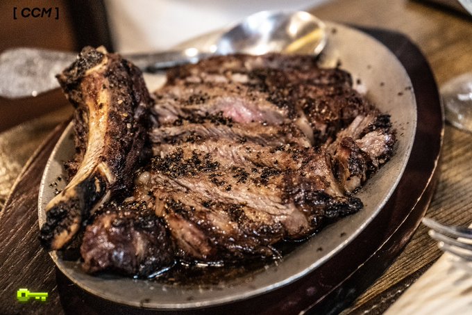 A massive, pepper-crusted tomahawk steak cooked medium-rare sits on a ceramic plate on a wooden board, served with a spoon in the background.