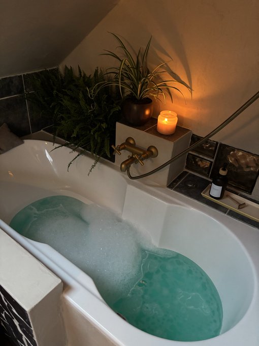 A white bathtub filled with bubbly turquoise water, surrounded by ferns, a potted plant, and a lit candle, creating a relaxing spa atmosphere.