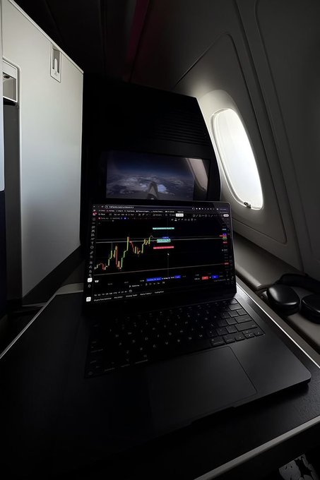 A laptop on an airplane tray table displays a trading chart with green and red candlesticks, positioned next to the window showing the wing in flight. The setting suggests a traveler multitasking between trading and air travel, with the destination teased in the post for guessing.