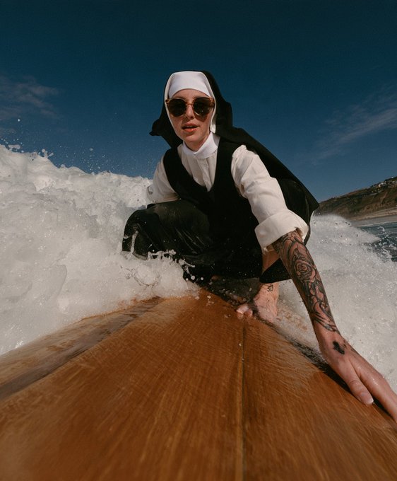A low-angle shot of a person wearing a nun habit and sunglasses surfing on a longboard while gripping the nose of the board.