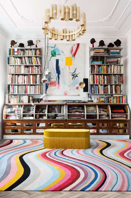 The image features a colorful, patterned rug with swirls of pink, blue, yellow, green, and red colors. The rug has a unique design that resembles a wave or a spiral shape. In front of the rug, there are several bookshelves filled with various books arranged in different sizes and orientations. A chandelier is also present above the rug, adding to the overall aesthetic appeal of the room. The post about irl being an art collector's favorite suggests that this rug might be a valuable item for someone interested in collecting unique and eye-catching home decor pieces.