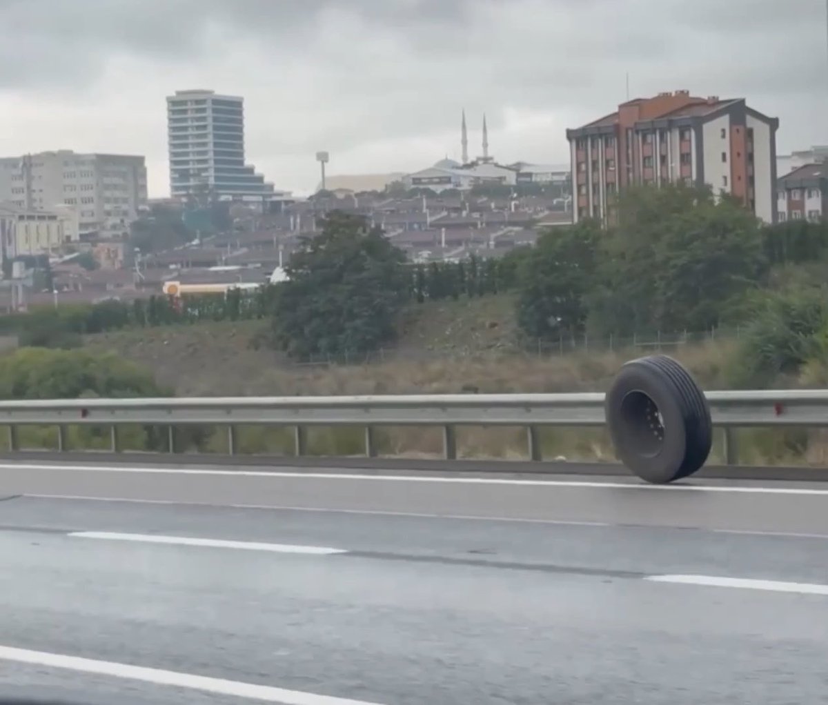 A photograph taken from a highway showing a tire rolling loose along a metal guardrail, with a city skyline featuring high-rise buildings and a mosque visible in the background.