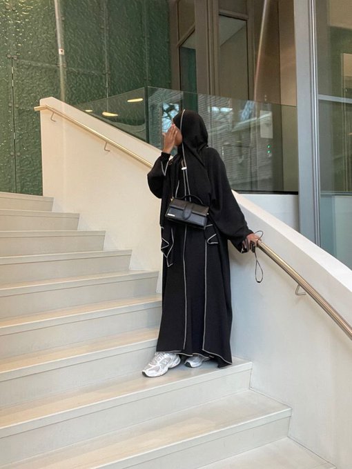 A person in a black hijab and traditional black robe with white trim standing on a staircase, holding a small black handbag.