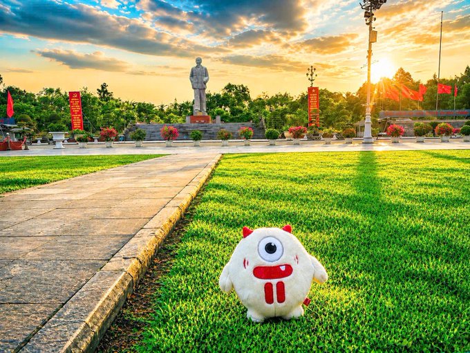 A small white one-eyed monster plush toy with red accents sits on green grass in a park, with a statue and sunset visible in the background.
