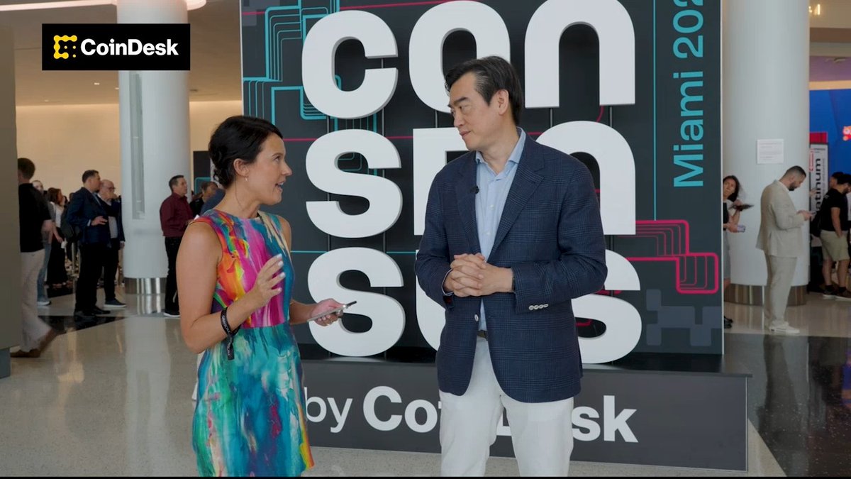 The image shows a man wearing a blue suit and a woman dressed in colorful clothing standing next to each other, possibly having a conversation or posing together. The backdrop of the scene features a large sign with the words "CoinDesk" written on it, indicating that they are at an event related to cryptocurrency or digital currency. The presence of multiple people around them suggests that this is a social gathering or a convention where attendees can network and discuss various topics, including the use of tokens for financial transactions in Asia.