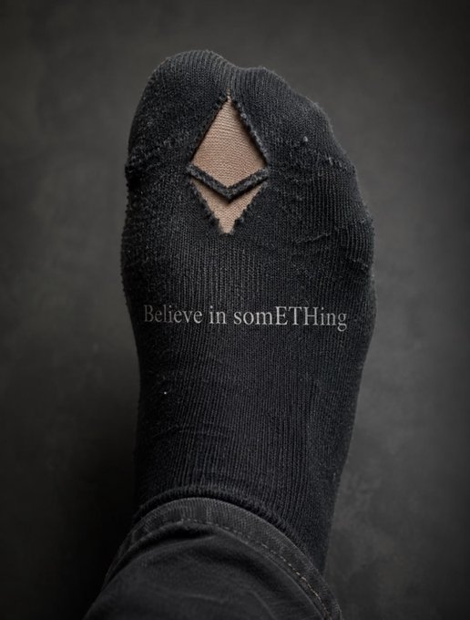 A worn black sock with a hole at the toe reveals skin underneath, displaying the text "Believe in somETHing" across its front, visually playing on the phrase's meaning of advocating for a cause.