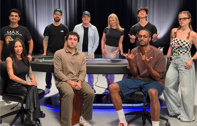 A group photo of eight people posed around a poker table in a studio setting, with a man seated in the center wearing a beige shirt and another gesturing in a brown shirt.