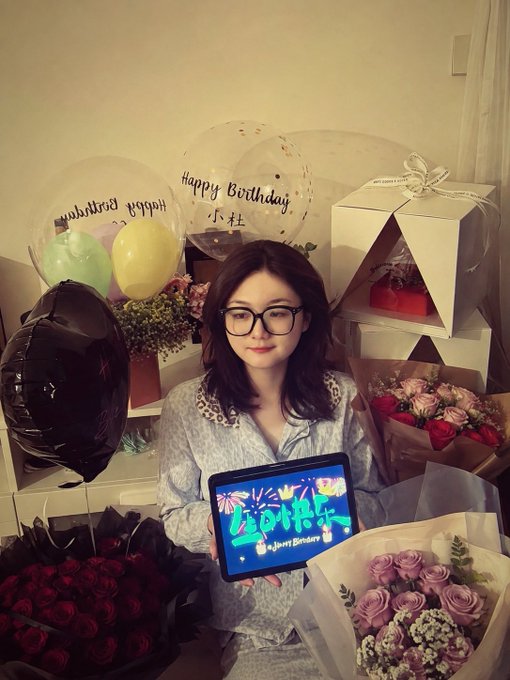 A woman wearing glasses sits surrounded by birthday decorations, including "Happy Birthday" balloons with the name "小杜" and a tablet displaying "Happy Birthday" graphics, alongside bouquets of red and pink roses and a black balloon. The celebratory scene contrasts humorously with the post's announcement of taking a birthday day off from work, with governance proposals jokingly replaced by a personal celebration.