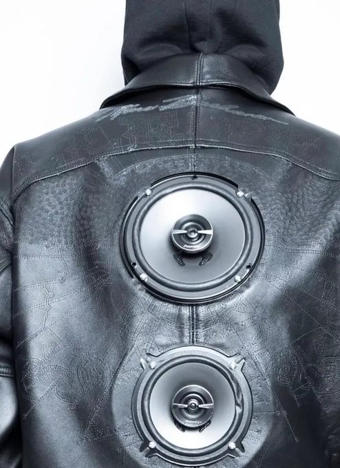 The back view of a person wearing a black leather hooded jacket featuring two large, round, metallic speaker-like fixtures attached to the back.