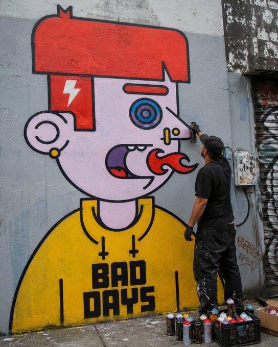 The image features a man painting graffiti on a wall while wearing a yellow shirt that says "Bad Days" and black pants. The man appears to be using spray paint cans as he creates his artwork. There are several cans of spray paint scattered around him, indicating an ongoing project or a completed piece. In the background, there is a building with graffiti art on it, suggesting that this area has been used for street art in the past.