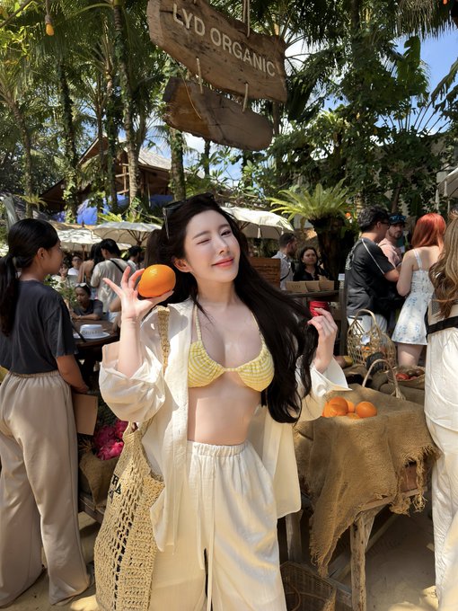 A woman in a yellow bikini top and white outfit posing with an orange at an outdoor market, with a wooden sign reading 'LYD ORGANIC' visible above her.