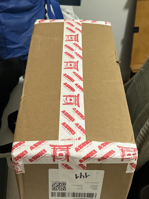 A cardboard shipping box sealed with red "Azuki" branded tape sits on a piece of furniture, featuring a white shipping label with a QR code and "447" handwritten in black marker. The package aligns with the post's mention of a newly delivered Azuki TCG order, with the collector expressing FOMO about being 2,830 miles away and unable to open it until Sunday.