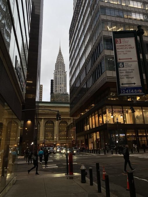 The image shows a busy city street with tall buildings lining both sides of the road. There are several people walking along the sidewalk, some carrying backpacks or handbags. A traffic light can be seen near the center of the scene, controlling the flow of vehicles and pedestrians. The sky above is cloudy, suggesting that it might be a cool evening in the city.