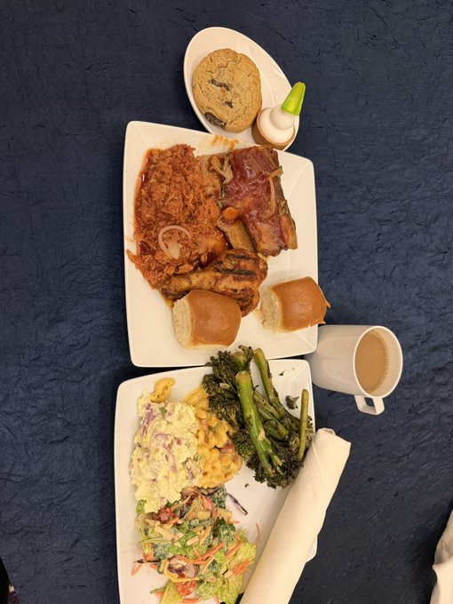 An overhead view of a meal spread on a dark table featuring a plate with fried chicken, bacon, sliders, a cookie, broccoli, macaroni salad, potato salad, and a cup of coffee.