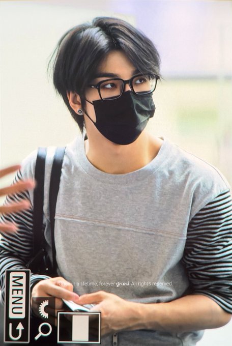 A young man with dark hair, wearing black-rimmed glasses, a black face mask, and a gray sweater with striped sleeves, is looking off to the side. The image includes interface elements like a "MENU" button, and a faint watermark reads "a lifetime, forever grew All rights reserved." The accompanying post about "XINLONG" and "ALPHADRIVEONE" appears unrelated to the visual content, suggesting this is a fan photo of a celebrity at an airport.