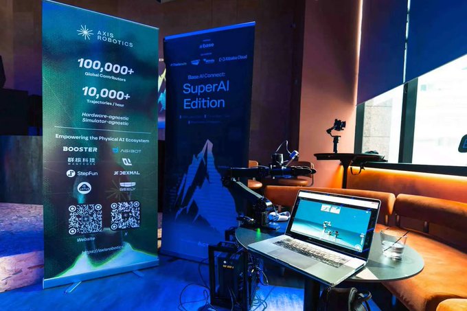 The image features a laptop computer sitting on top of a table, with a banner next to it that says "Physical AI onchain isn'qe uite accessible to everyone". The banner also mentions that there's an event happening at SuperAI headquarters in San Francisco. A person is standing behind the table holding a camera and another person is sitting nearby. There are two chairs visible in the scene, one near the laptop and the other further away from the table.