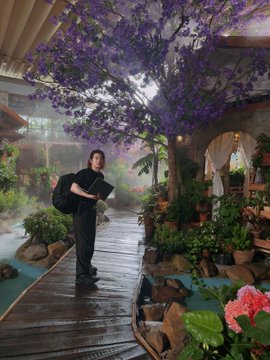 A person with a backpack stands on a wet wooden walkway in a lush indoor garden, holding a book while purple jacaranda flowers cascade from above.