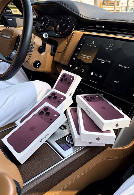 Multiple unopened boxes of purple/burgundy colored iPhone 17 Pro boxes are stacked on the passenger seat of a luxury vehicle, with the dashboard navigation screen displaying 'Sel Houston' and 'Muhammad's Phone'.