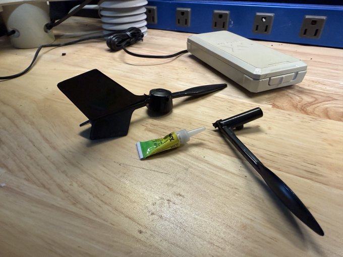 Black plastic components, possibly model airplane parts, rest on a wooden workbench alongside a small tube of adhesive and a white case, with a blue power strip in the background.