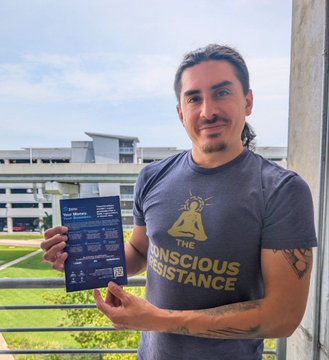 A man wearing a 'The Conscious Resistance' t-shirt stands on a balcony holding a blue informational flyer with a campus building in the background.