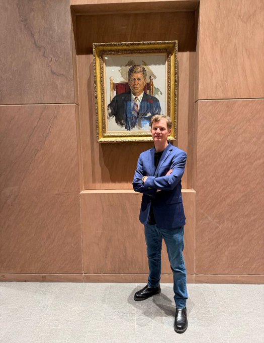 A man wearing a blue blazer stands with arms crossed in front of a framed portrait hanging on a wood-paneled wall.