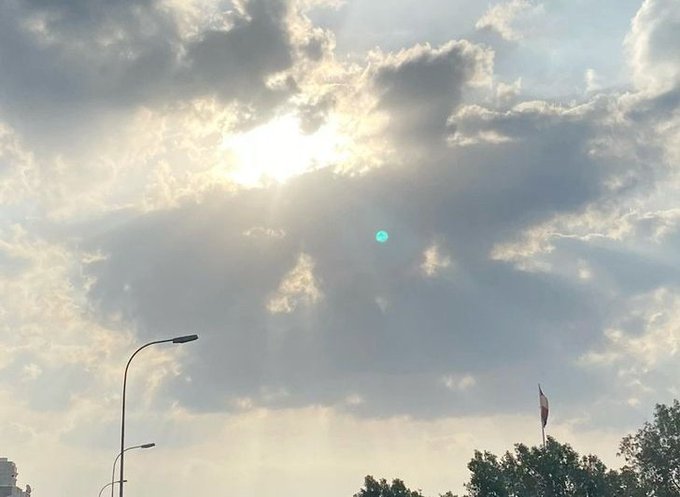 The image features a vibrant sky with an upside-down blue sun peeking through some white clouds. The sun's rays are shining directly onto the clouds, creating interesting shadows and highlights across the sky. In the distance, there is a street lamp standing tall against the backdrop of this beautiful scene.