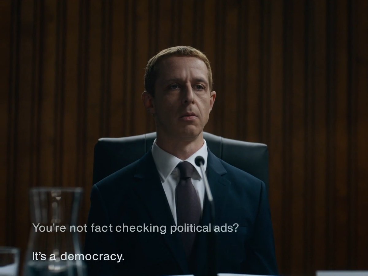 A serious-looking man in a dark suit and tie sitting at a desk with a microphone, with subtitle text reading 'You're not fact checking political ads? It's a democracy.'