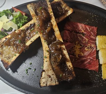 A plated meal featuring bone marrow on cross-cut bones topped with sauce, accompanied by slices of rare steak and yellow pickled vegetables on a black plate.