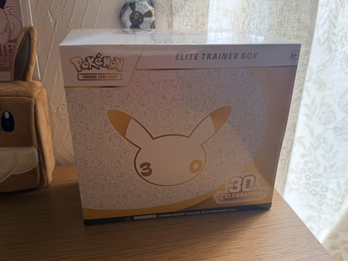 A Pokemon Trading Card Game Elite Trainer Box celebrating 30 years, featuring a gold-toned design with a stylized Eevee silhouette and patterned background, sitting on a wooden surface next to an Eevee plush.