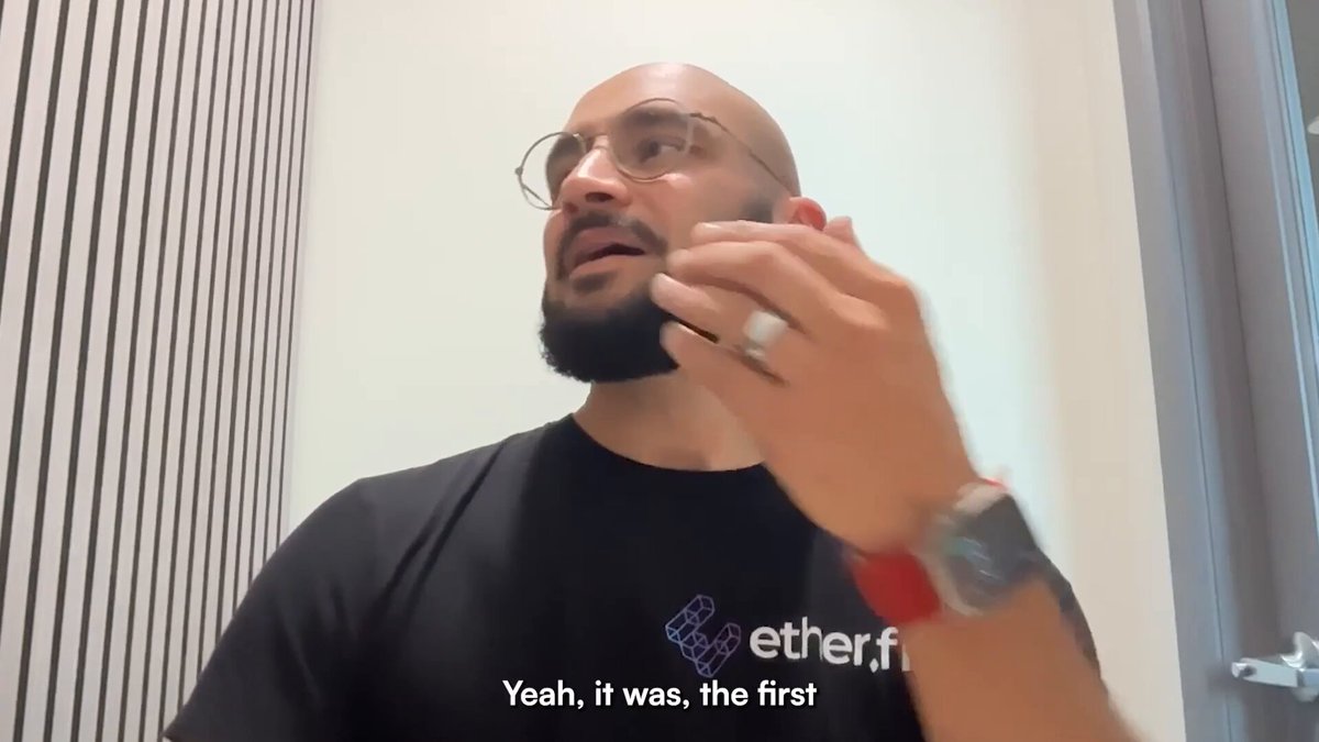 The image shows a man wearing glasses standing against a white wall with a door nearby. He appears to be talking on his cell phone while holding up his hand for emphasis, possibly discussing something important or sharing an interesting piece of information related to the post about DeFi and decentralized theater that he is viewing. The scene captures the essence of modern communication technology and its impact on various aspects of our lives.