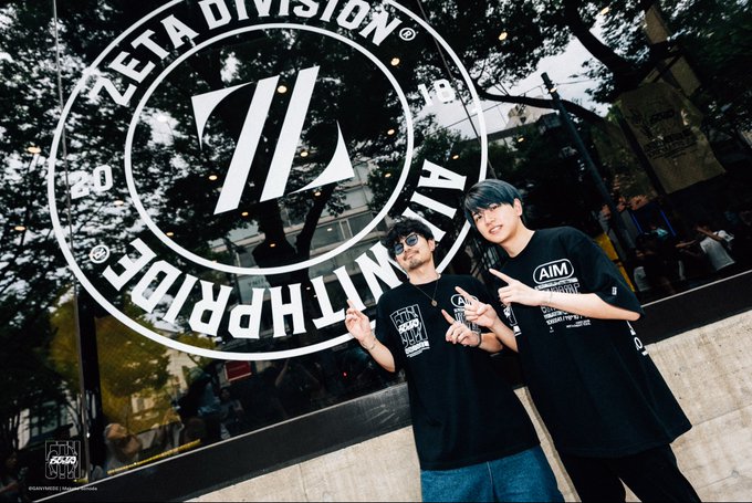 Two young fans pose in matching black "ZETA DIVISION" t-shirts, flashing peace signs in front of a large circular "ZETA DIVISION ALLMULTPRIDE" logo display, commemorating their visit to the brand's 5th anniversary pop-up store.