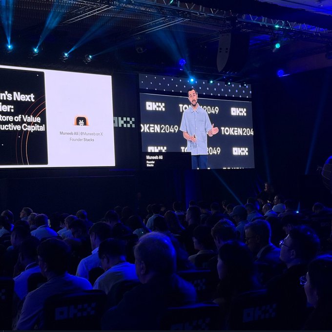 A photograph taken from the audience at the TOKEN2049 conference showing a presenter on stage with 'OKX' and 'TOKEN2049' branding, with the speaker identified as 'Muneeb Ali | @Muneeb on X' from 'Stacks'.