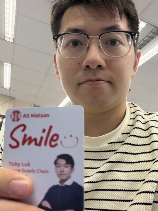 A young man with glasses holds up an "AS Watson" employee ID badge that reads "Smile" and identifies him as "Toby Luk."