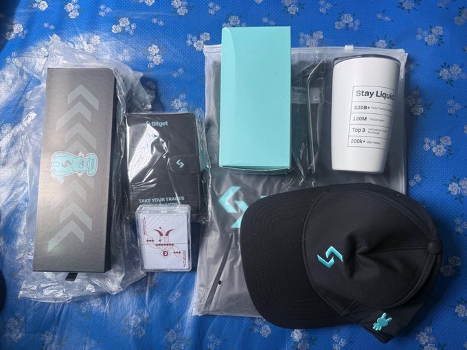 A collection of Bitget merchandise including a black box with arrows, branded cards, a 'Stay Liquid' tumbler with specifications (520B+, 120M, Top3, 200K+), and a black cap with the Bitget logo laid out on a floral blue surface.