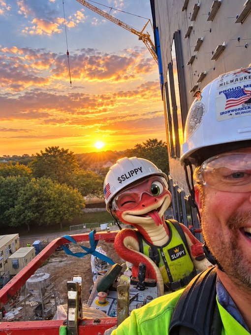 A selfie of a construction worker wearing a white hard hat with 'MILWAUKEE' and 'IRONWORKERS' stickers, alongside a snake character figurine wearing a hard hat labeled 'SLIPPY', against a sunset sky with a crane.