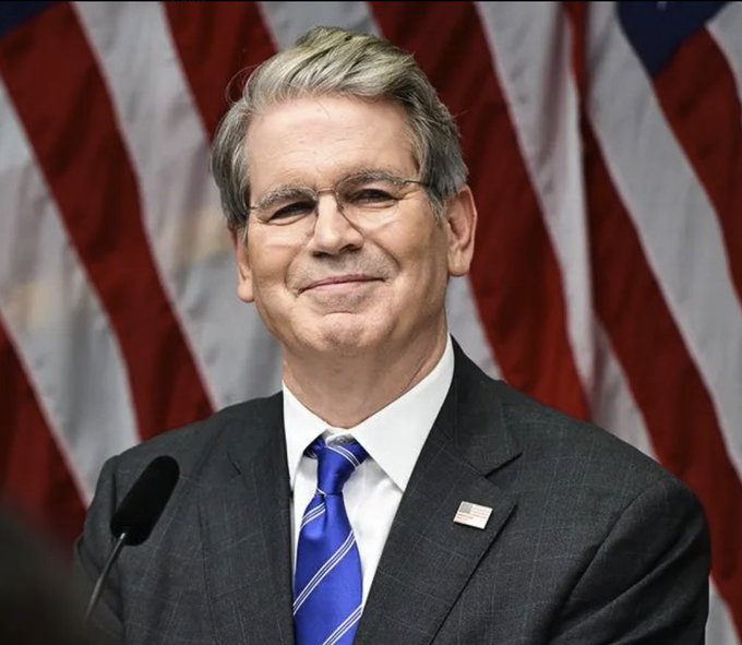 Scott Bessent, the U.S. Treasury Secretary, stands at a podium with a microphone in front of an American flag backdrop, smiling slightly while wearing a dark suit, blue striped tie, and American flag lapel pin. The image accompanies news that Bessent could deploy nearly $1 trillion from the Treasury General Account to fund bond buybacks, a move framed as bullish for the crypto market, particularly Solana ($SOL).